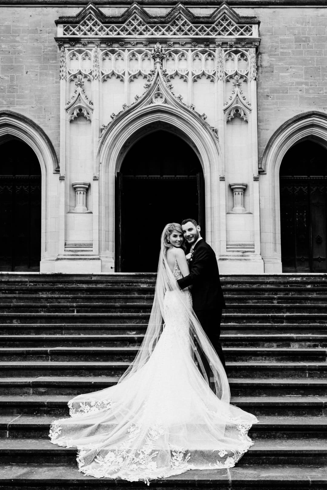 A couple embraces on a grand staircase in front of ornate architectural details, showcasing their love on their special day.