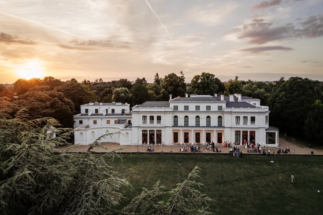 Aerial view of Gunnersbury Park surrounded by lush greenery, with wedding guests mingling outside and a beautiful sunset in the background.