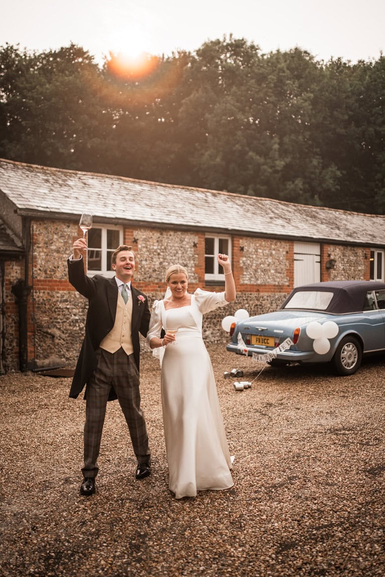Enjoy a joyful wedding moment capturing bride and groom celebrating with champagne beside vintage convertible, outdoor country wedding photos by Alistair Jones.